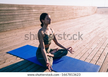 From above concentrated young slim woman in sportswear and with black hair sitting on mat practicing yoga in lotus pose keeping calm and closed eyes in wooden promenade on background of sunlight 