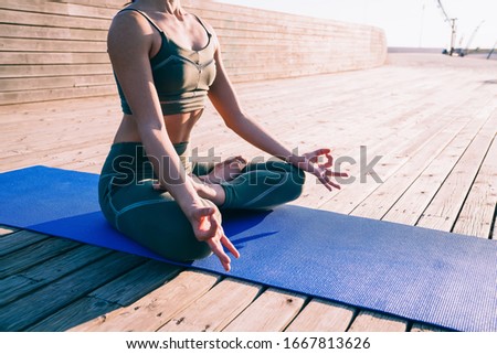 Crop faceless sporty slim female in tracksuit sitting on mat practicing yoga in lotus pose in wooden promenade on background of sunlight having healthy life 