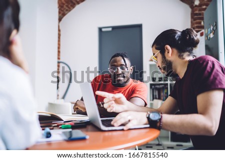 Young male freelance colleagues in casual clothes working with laptop and discussing website design while sitting at table during business meeting