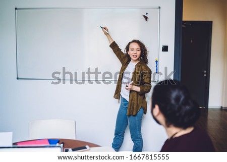 Motivated youthful woman in casual clothes writing business information on wall board and talking to colleagues while having meeting in office
