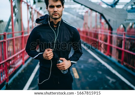 Sporty man with muscular body listening music podcast while running across city bridge, young jogger doing cardio exercises and workout outdoors making effort for achievement of wellness goal