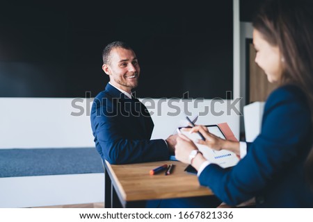 Back view from female boss using modern digital tablet for banking payment during cooperation meeting with cheerful man sitting in front and smiling, successful professionals enjoying brainstorming