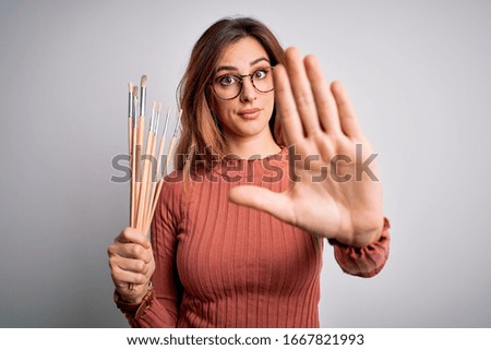 Young beautiful brunette artist woman holding painter brushes over white background with open hand doing stop sign with serious and confident expression, defense gesture