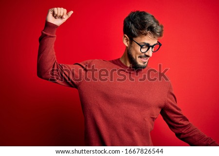 Young handsome man with beard wearing glasses and sweater standing over red background Dancing happy and cheerful, smiling moving casual and confident listening to music