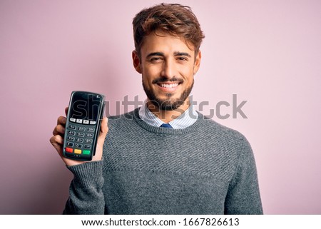 Young man paying using dataphone standing over isolated white background with a happy face standing and smiling with a confident smile showing teeth