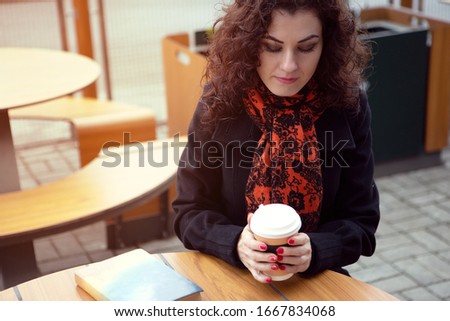 A young woman in a coat and colorful wool scarf is drinking coffee while reading a book on the terrace. Photo with artistic noise