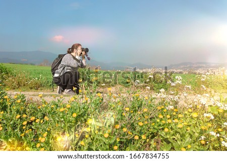 Young woman with a camera in a meadow taking nature photos at day time. Nature traveller against a blue sky with empty copy space for Editor's text.