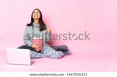 Young mixed race woman eating popcorn while watching a movie on the laptop pointing up and surprised