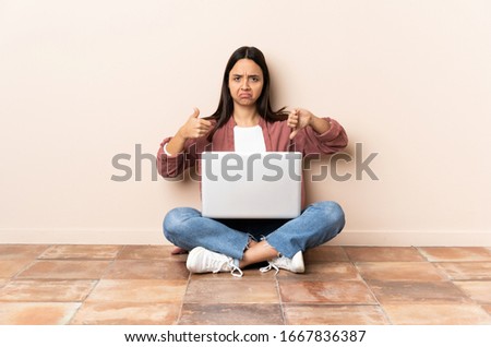Young mixed race woman with a laptop sitting on the floor making good-bad sign. Undecided between yes or not