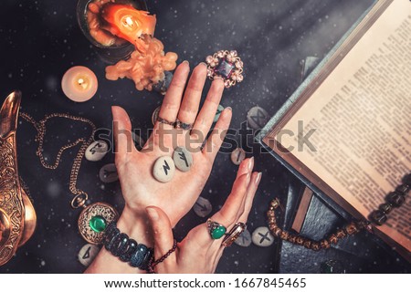 Astrology and esotericism. The witch is holding in his hands the rune stones. On a black background lie fortune-telling runes, a book, precious amulets, a copper lamp and a candle. Dust and light Royalty-Free Stock Photo #1667845465
