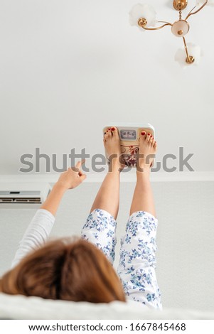 A woman is lying on a bed with her legs raised and electronic scales lying on them. Bottom view. The concept of weight loss and weight gain.