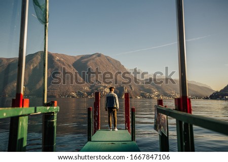 Man standing on a pier on the lake Lugano in Switzerland. Travel lifestyle concept. Traveler looking at the mountains, standing on a wooden green pier. Back view.