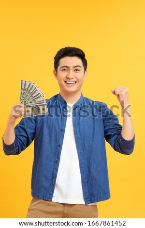 Successful Asian man. Happy young man holding money while standing and arms up, isolated on yellow background