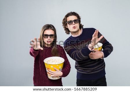 Young shocked couple, woman and man in 3d glasses watching movie film on date, holding bucket of popcorn, pointing index fingers on copyspace isolated on white background.