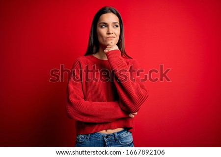 Beautiful young brunette woman wearing casual sweater standing over red isolated background with hand on chin thinking about question, pensive expression. Smiling with thoughtful face. Doubt concept.