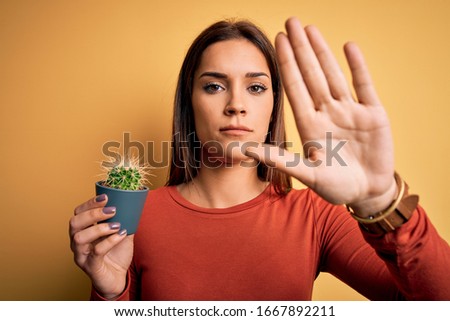 Young beautiful brunette woman holding small cactus pot over yellow background with open hand doing stop sign with serious and confident expression, defense gesture