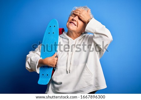 Senior beautiful sporty woman holding skate standing over isolated blue background stressed with hand on head, shocked with shame and surprise face, angry and frustrated. Fear and upset for mistake.