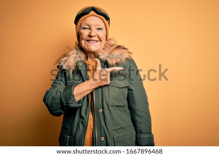 Senior beautiful grey-haired skier woman wearing snow sportswear and ski goggles cheerful with a smile of face pointing with hand and finger up to the side with happy and natural expression on face