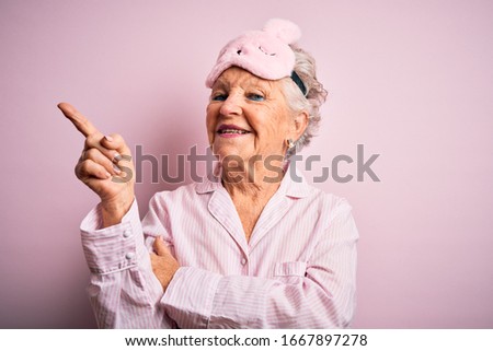 Senior beautiful woman wearing sleep mask and pajama over isolated pink background with a big smile on face, pointing with hand and finger to the side looking at the camera.