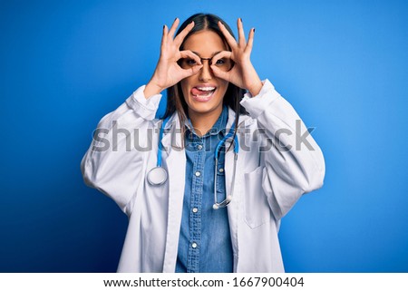 Young beautiful doctor woman wearing stethoscope and glasses over blue background doing ok gesture like binoculars sticking tongue out, eyes looking through fingers. Crazy expression.