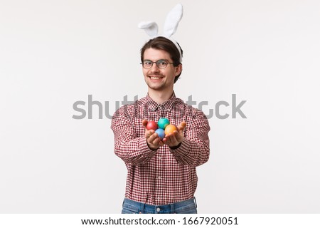 Traditions, religious holidays, celebration concept. Happy kind and cute european man in rabbit ears, glasses, giving painted eggs for Easter day, smiling heartwarming, stand white background
