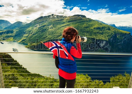 Tourism holidays picture and traveling. Woman tourist enjoying fjord landscape Geirangerfjord from Ornesvingen eagle road viewpoint, taking photo with camera, Norway.