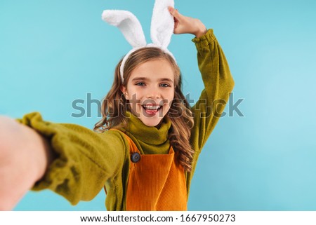 Image of a cute positive young girl child in easter holiday bunny ears take a selfie by camera isolated over blue wall background.