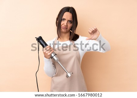 Young brunette girl using hand blender over isolated background showing thumb down