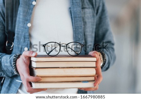 Close up view of ,ale young student in jeans clothes that is in corridor of a college with books with eyewear on it in hands.