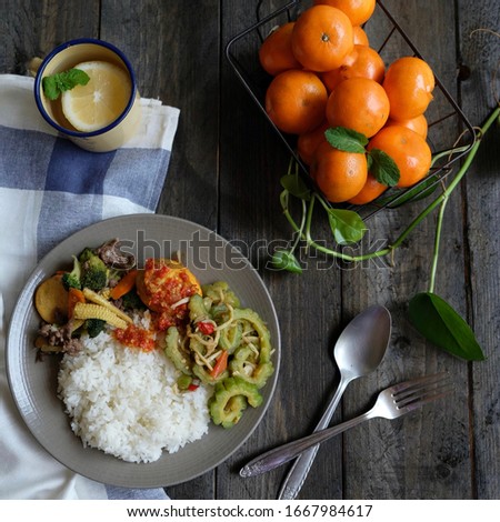 Flat-lay concept for food photography,  plate of lunch sapo tahu, telur balado and bitter melon stir fry. A basket of oranges, enamel cup, spoon and fork on black wooden background.