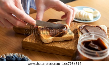 Spreading chocolate nut butter on toasted bread. Female hands smear chocolate spread on sandwich bread. Preparing lunch or breakfast Royalty-Free Stock Photo #1667986519