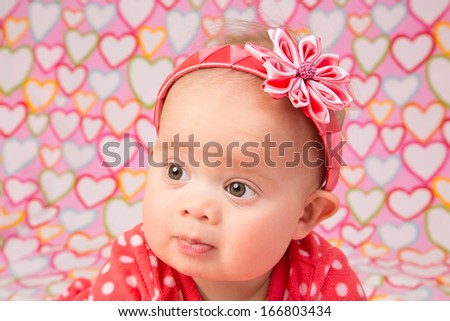 An adorable baby girl wearing a red headband with a decorative flower, lying on a blanket with hearts design printed on it