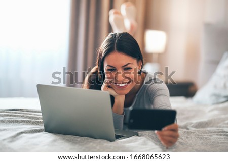 A beautiful girl lies in bed next to laptop and posing while taking a selfie with her phone.