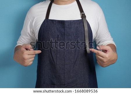 Portrait of male Asian chef or waiter pointing showing his apron, proud confident chef concept, against blue background with copy space