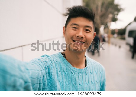 Portrait of young Asian man looking confident and taking a selfie while standing outdoors in the street. Royalty-Free Stock Photo #1668093679