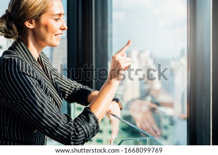 Portrait of businesswoman wearing black and white suit points finger to transparent at glass window modern city background. With copy space for your text.