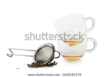 Two glass cups of drunk green tea stacked and a tea strainer of stainless steel with sprinkled some green tea on a white background