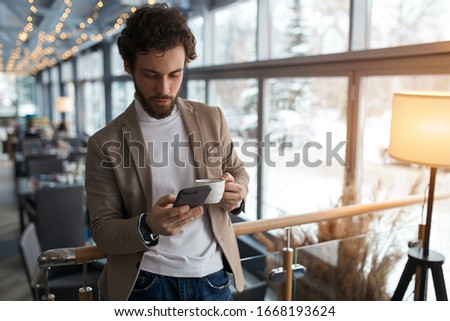 young man sending message to colleague. confident attractive young man holding smart phone and looking at it while standing in front of the big window in cafe, close up photo, free time, spare time Royalty-Free Stock Photo #1668193624