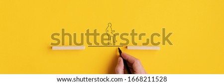 Wide view image of male hand drawing a line between two wooden pegs for a silhouette of a man to walk across. Over yellow background with copy space. Royalty-Free Stock Photo #1668211528