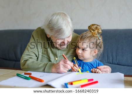 Beautiful toddler girl and grand grandmother drawing together pictures with felt pens at home. Cute child and senior woman having fun together. Happy family indoors