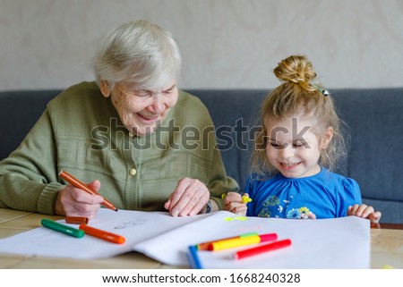 Beautiful toddler girl and grand grandmother drawing together pictures with felt pens at home. Cute child and senior woman having fun together. Happy family indoors
