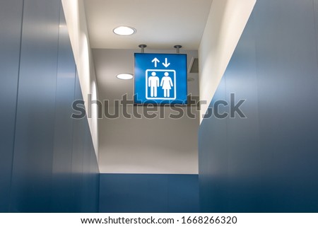 An illuminated sign with the icon of a lift hang on ceiling of the blue hallway. Direction way for a public elevator at corridor.