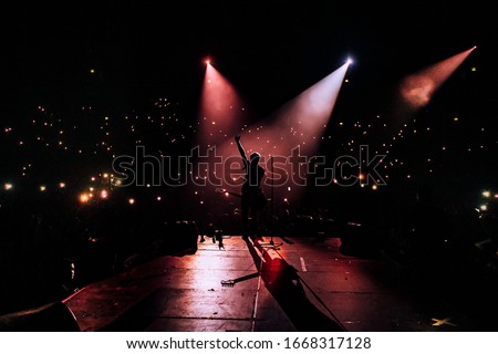 Music show. Guitarist in front of crowd on scene in night club. Bright stage lighting, crowded dance floor. Phone lights at concert. Band red silhouette crowd. People with cell phone 