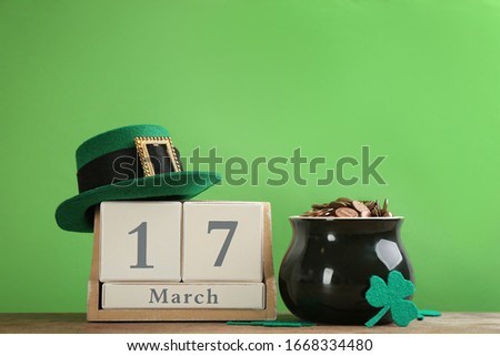 Composition with pot of gold coins and wooden block calendar on table against green background. St. Patrick's Day celebration