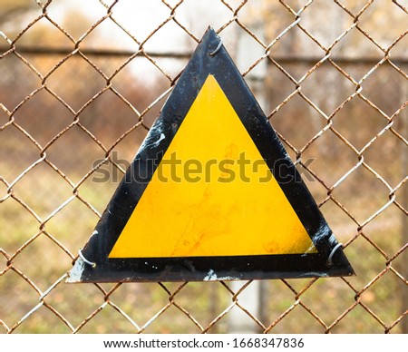 Triangle warning sign on the metal mesh fence. Basic warning sign triangle with a black border and amber background.