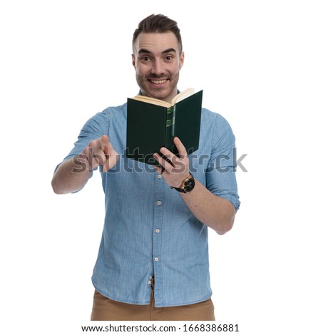 Happy casual holding book and pointing, laughing while wearing blue shirt, standing on white studio background