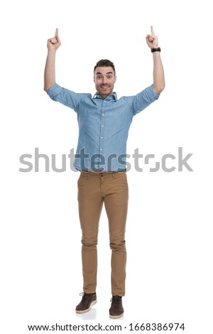 Happy casual man pointing up with both hands while wearing blue shirt, standing on white studio background