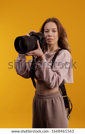 Young girl freelancer photographer posing with photo camera in hands on a yellow background