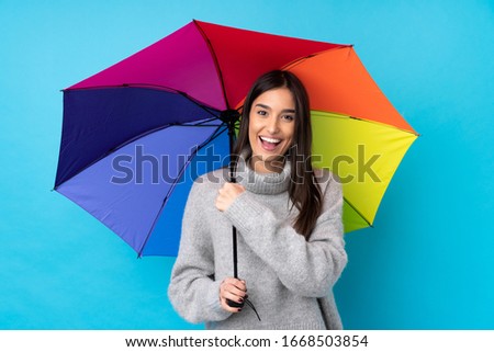 Young brunette woman holding an umbrella over isolated blue wall celebrating a victory