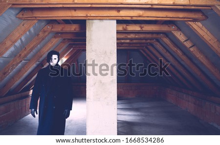 A scary guy in white mask and long dark coat standing in an abandoned attic. Scary and mystical picture. Horror concept.